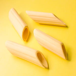 May include: Four penne pasta shapes, light in colour, are displayed against a bright yellow backdrop. The pasta has a natural, wooden look with visible grain and open ends, suggesting a rustic or handmade quality.