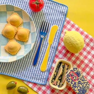 May include: A still life featuring a plate of ravioli, a red tomato, green olives, a yellow lemon, and a sardine can. A blue and yellow fork and knife are on a blue and white striped napkin. The background is yellow and red checkered.