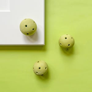 May include: Three pale green, spherical cabinet knobs with multiple small holes. One knob is attached to a white cabinet door, while the other two are on a bright green background. The knobs have a matte finish.