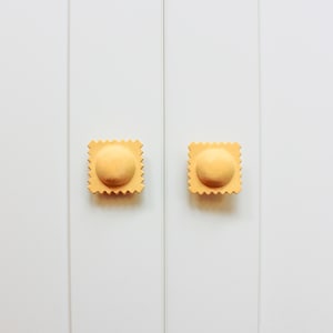 May include: Two yellow cabinet knobs shaped like ravioli pasta. The knobs are mounted on a white cabinet door.