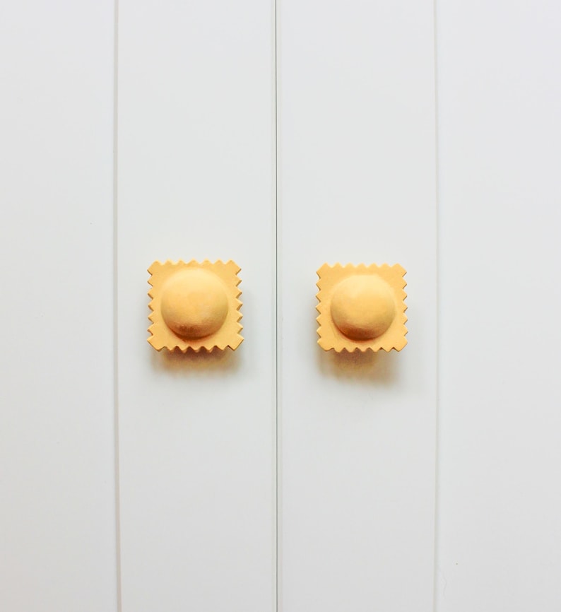 May include: Two yellow cabinet knobs shaped like ravioli pasta. The knobs are mounted on a white cabinet door.