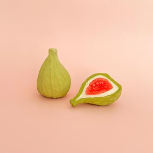 May include: Two fig sculptures on a pink background. One whole fig and one fig cut in half, revealing a bright red interior with a white border. The figs are green with textured surfaces.