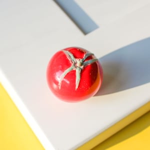 May include: A glossy, red tomato figurine with green accents on the stem. The tomato is set against a white surface with a yellow background, creating a vibrant contrast. The figurine is a decorative item.