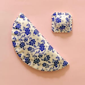May include: Two ceramic items with a blue floral design on a cream base. One is a half-moon shape, the other a square with a scalloped edge. The items are set against a pink background.