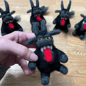 May include: Five black felt toys shaped like horned devils with red tongues sticking out. The toys are sitting on a wooden surface.