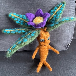 Könnte beinhalten: Ein fantasievolles Filzspielzeug, das eine Pflanze mit einer lila Blume und grünen Blättern mit blauen Akzenten darstellt. Die Pflanze hat einen braunen Filzkörper mit einem Gesicht und Armen.