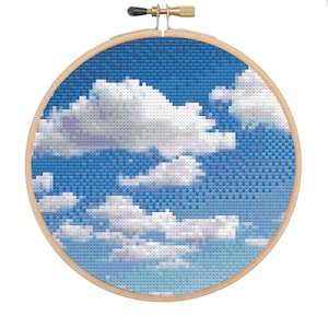Pode incluir: Um aro de bordado redondo com um desenho de ponto cruz de um céu azul com nuvens brancas. O céu é azul claro com um degradê de azul mais escuro no topo. As nuvens são brancas e fofas, e estão dispostas em um padrão aleatório no céu.