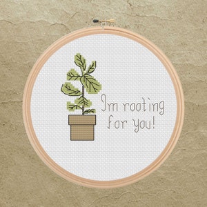 May include: Cross-stitch embroidery in a wooden hoop. The design features a potted plant with green leaves and the text "I'm rooting for you!" in a simple font. The background is a textured, tan color.