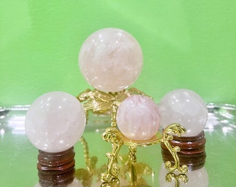 Könnte beinhalten: Vier Kristallkugeln in verschiedenen Rosa- und Weißtönen. Eine große Kugel steht auf einem goldfarbenen Dekorationsständer, die anderen auf Holzsockeln und einem kleineren Goldständer. Der Hintergrund ist einfarbig grün.