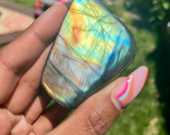 May include: A smooth, iridescent labradorite gemstone with a blue, green, and gold sheen. The stone is held in a hand with colorful nail polish.