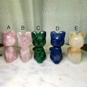May include: Five carved crystal cat figurines in different colors. The cats are sitting with their tails curled around their legs. From left to right, the colors are pink, white, green, blue, and yellow.