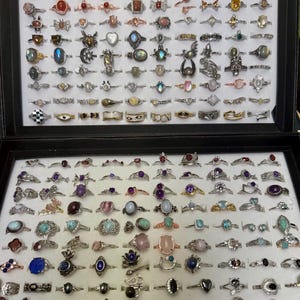 Puede incluir: Una colección de anillos de plata con diversas piedras preciosas, incluyendo amatista, turquesa, piedra lunar y cuarzo rosa. Los anillos se muestran en una caja de terciopelo negro.