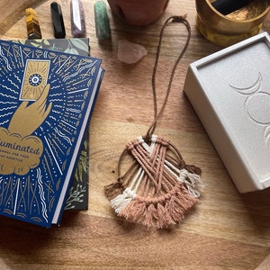 May include: A blue book titled "Illuminated: A Journal for Your Tarot Practice" is on a wooden surface with a white box featuring a triple moon symbol, a small macrame dreamcatcher, and various crystals.