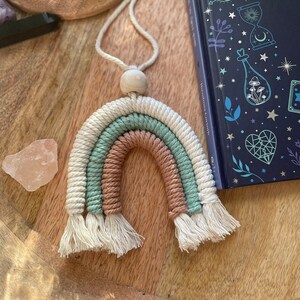 Może przedstawiać: Wisząca dekoracja w kształcie tęczy wykonana techniką makramy w jasnobrązowych, jasnozielonych i białych odcieniach. Tęcza jest wykonana z węzłowatego sznurka i ma drewnianą koralik na górze.