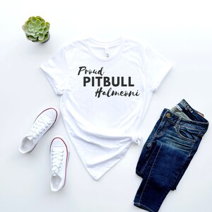 May include: A white t-shirt with the text "Proud PITBULL Halmeoni" in black script. The shirt is paired with blue jeans and white canvas sneakers with red accents. A small green succulent plant is also visible.