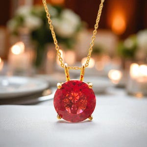May include: A gold-colored necklace featuring a large, round, faceted red gemstone pendant. The pendant is held in place by four gold prongs. The chain is delicate and also gold-colored. The background is blurred, suggesting a soft focus.