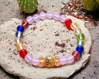 May include: A beaded bracelet with a mix of colorful glass beads and a gold-colored centerpiece. The bracelet features pink, red, yellow, green, and blue beads. The bracelet is resting on a light-colored stone.