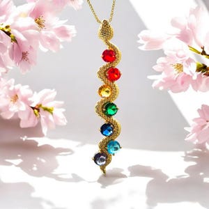 May include: A gold-colored snake pendant necklace with seven colorful gemstones. The gemstones are red, orange, yellow, green, blue, indigo, and violet. The necklace is displayed against a white background with pink flowers.