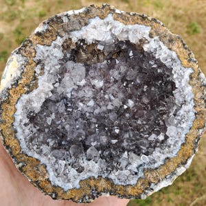 May include: A circular amethyst geode featuring a deep purple crystalline center surrounded by a white quartz layer and a rough, textured brown outer crust. The interior is filled with numerous small, pointed, translucent purple crystals.