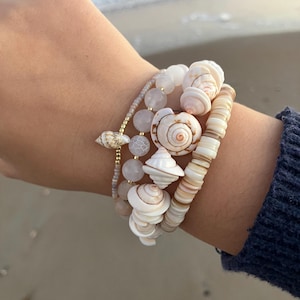 May include: A collection of bracelets featuring natural seashells and beads. The bracelets include white, tan, and pink elements, with a focus on the unique shapes and textures of the seashells. The bracelets are stacked on a wrist.