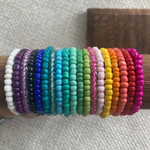 May include: A stack of 20 colorful beaded bracelets in a rainbow pattern. The bracelets are made of small, round beads in various shades of purple, blue, green, yellow, orange, and red.
