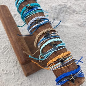 Könnte beinhalten: Eine Sammlung von geschichteten Kordelarmbändern auf einem Holzständer. Jedes Armband hat einen dunklen Haifischzahn-Anhänger, der mit goldfarbenem Draht umwickelt ist. Die Kordeln variieren in Blau-, Hellblau-, Weiß-, Beige- und Orangetönen.