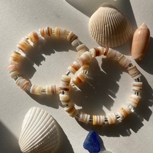 Op de afbeelding: Twee armbanden gemaakt van gelaagde schelpen met zilveren accenten. De armbanden liggen op een wit oppervlak met schelpen en zeeglas.