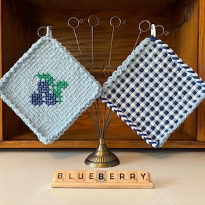 May include: Two square potholders hanging on a metal stand. One is light blue with a dark blue and green cross-stitched blueberry design. The other features a dark blue and white gingham check pattern. A wooden tile rack displays the word BLUEBERRY.