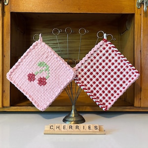 May include: Two handmade pot holders. One is light pink with a cherry design, the other is red and white checkered. The word "CHERRIES" is spelled out with letter tiles. Kitchen decor.