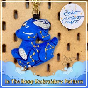 May include: A blue plush toy with gold embroidery of constellations. The toy is attached to a gold key chain and is hanging on a pegboard. The text "Pocket Artist Crafts" is visible on a sticker in the upper right corner of the image.