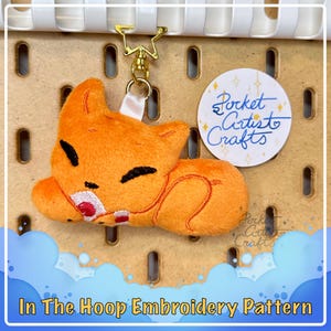 May include: Orange plush fox keychain with embroidered details, sleeping with a red heart in its paws. The keychain is attached to a gold chain and a white ribbon. The image includes the text "Pocket Artist Crafts".