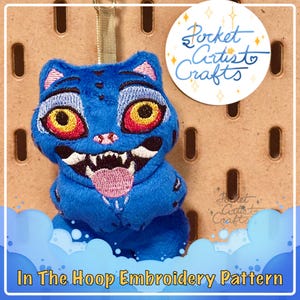 May include: A blue plush cat with embroidered details, including large yellow and red eyes, a pink tongue, and sharp white teeth. The cat is hanging from a gold ribbon. The image includes the text "Pocket Artist Crafts" and "In The Hoop Embroidery Pattern."