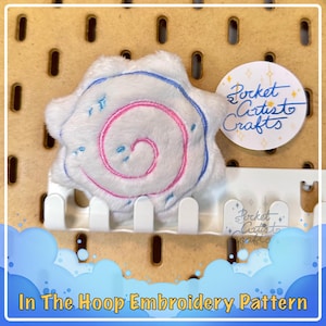Op de afbeelding: Een witte pluche swirl met roze en blauwe borduurwerk, bevestigd aan een prikbord met een witte plastic prikbordhaak. De swirl is een digitaal borduurontwerp voor machineborduurwerk. Het ontwerp is gelabeld "Pocket Artist Crafts" in een cirkel met een witte achtergrond en gouden glitter.
