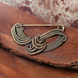 May include: A gold wire brooch with a unique, abstract design. The brooch is made of wire that has been woven together to create a intricate pattern.