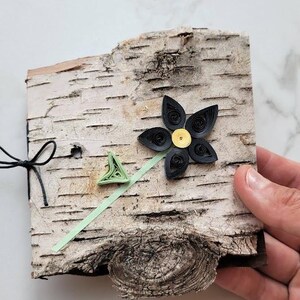 May include: A handmade birch bark clock with a black paper flower and a yellow center. The clock has a light green paper arrow and a black bow. The clock is held by a hand.