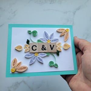 May include: A rectangular greeting card with a teal border and white center featuring paper quilling art. Three wooden tiles in the middle display the letters C & V. The design includes light blue, peach, and green paper coils shaped into flowers and leaves.