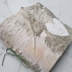 May include: A square piece of birch bark with a white heart-shaped cutout affixed to the surface. A green string is tied in a bow on the left side. The bark has a natural, textured appearance with shades of gray, white, and tan.