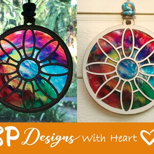 K&ouml;nnte beinhalten: Ein runder, bunter Holzschmuck mit einem Buntglasdesign. Der Anh&auml;nger zeigt eine Blume mit einem blauen Zentrum und ist von einem schwarzen Rahmen umgeben. Der Anh&auml;nger h&auml;ngt an einer Schnur.