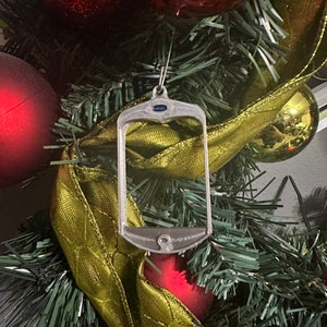 May include: A silver-colored Christmas ornament shaped like a cell phone with a blue jewel at the top. The ornament is hanging on a green artificial Christmas tree with red and gold ribbon.