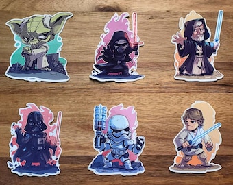 May include: Six colorful stickers featuring characters from Star Wars. The stickers are on a wooden surface. The characters include Yoda, Darth Vader, Kylo Ren, Obi-Wan Kenobi, Luke Skywalker, and a Stormtrooper.