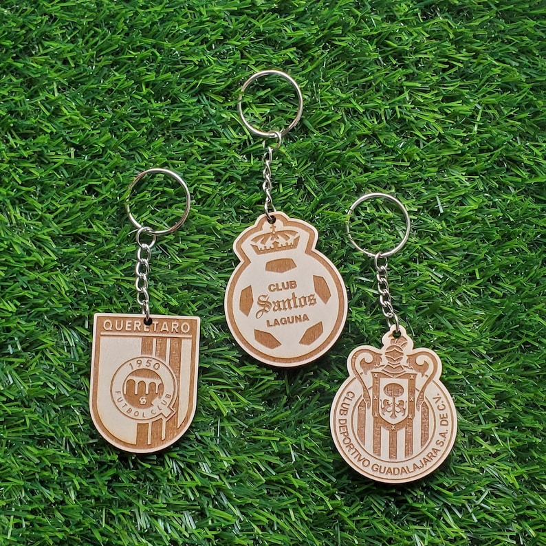 May include: Three wooden keychains with different soccer team logos. The keychains are brown and feature the logos of Club Santos Laguna, Club Deportivo Guadalajara, and Queretaro Futbol Club.
