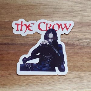 May include: A white sticker featuring a black and white illustration of a character from the film "The Crow". The title "The Crow" is in red text above the character.