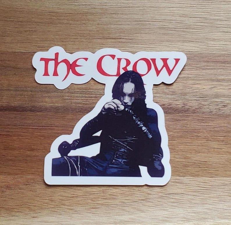 May include: A white sticker featuring a black and white illustration of a character from the film "The Crow". The title "The Crow" is in red text above the character.