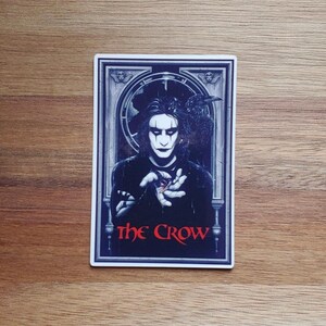 May include: A sticker featuring a black and white illustration of a man with a crow on his shoulder. The man is holding a red heart in his hand. The text "The Crow" is written in red letters at the bottom of the sticker.