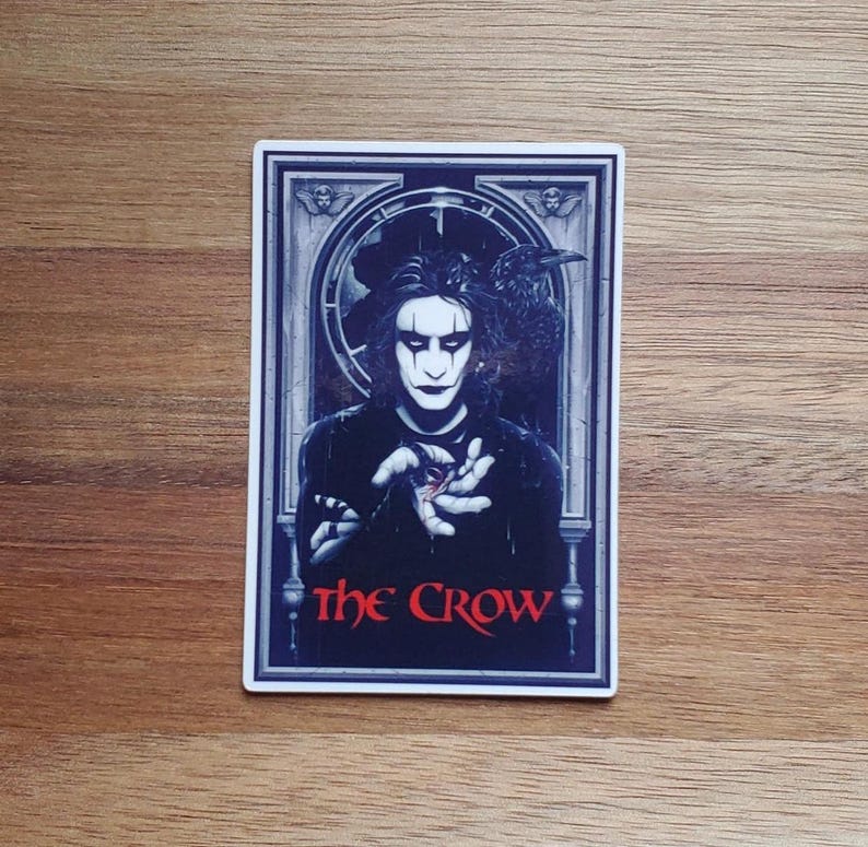 May include: A sticker featuring a black and white illustration of a man with a crow on his shoulder. The man is holding a red heart in his hand. The text "The Crow" is written in red letters at the bottom of the sticker.