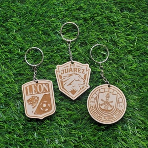 May include: Three wooden keychains with different soccer team logos. The keychains are on a green grass background. The logos are for FC Juarez, Leon FC, and Mazatlan Futbol Club.
