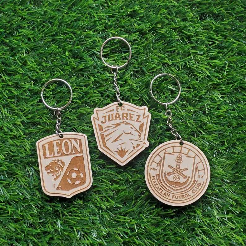 May include: Three wooden keychains with different soccer team logos. The keychains are on a green grass background. The logos are for FC Juarez, Leon FC, and Mazatlan Futbol Club.