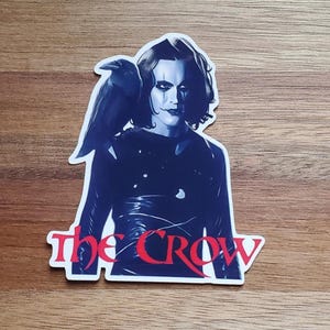 May include: A blue and white sticker featuring a character from the film "The Crow" with a crow on his shoulder. The text "The Crow" is in red letters.