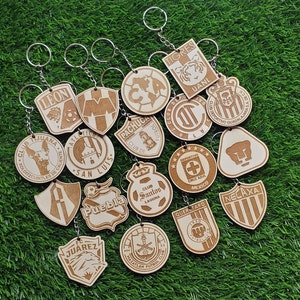 May include: A collection of 16 wooden keychains featuring logos of different Mexican soccer teams. The keychains are made of light brown wood and have a variety of designs, including team names, crests, and mascots.