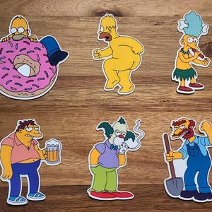 May include: A set of six stickers featuring various Simpsons characters. The stickers are printed on white paper with a glossy finish. The characters include Homer, Marge, Bart, Lisa, Maggie, and Krusty the Clown.